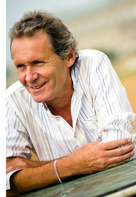 A smiling man with short, tousled hair, wearing a white striped shirt, leaning on a surface outdoors.