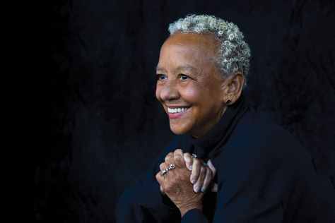 Smiling elderly woman with short curly gray hair, wearing a black top, sitting against a dark backdrop.